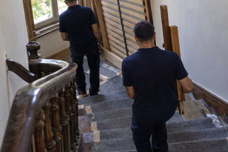 Déménageurs qui portent un sommier et les montants d'un lit démonté, escalier ancien en bois aux marches couvertes de protections