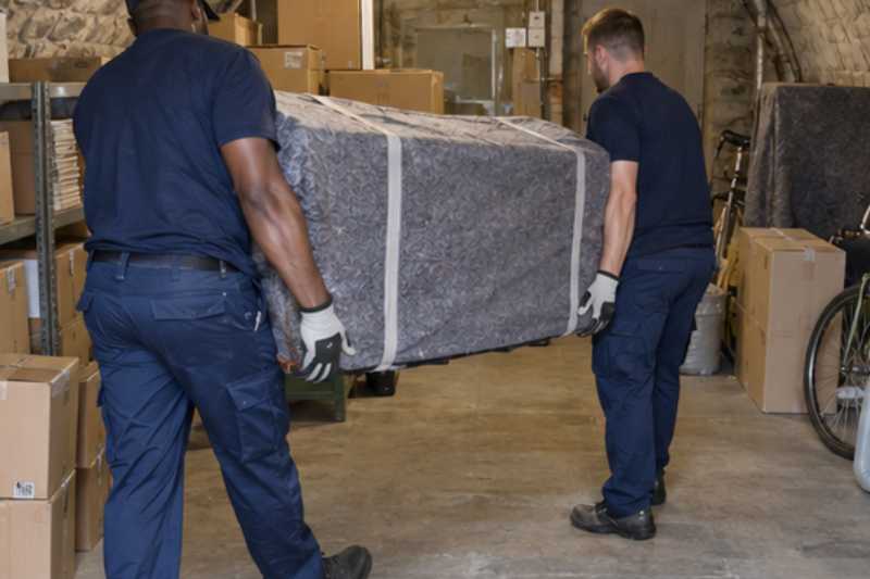 Deux déménageurs portent des meubles emballés sous couvertures, cave voûtée d'immeuble avec cartons