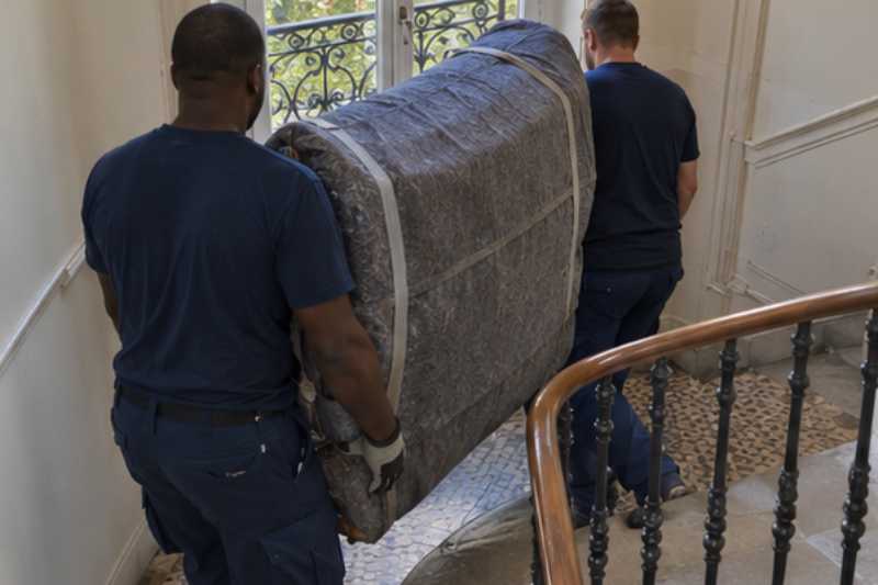 Deux déménageurs portent des meubles emballés sous couvertures, escalier tournant avec un palier étroit