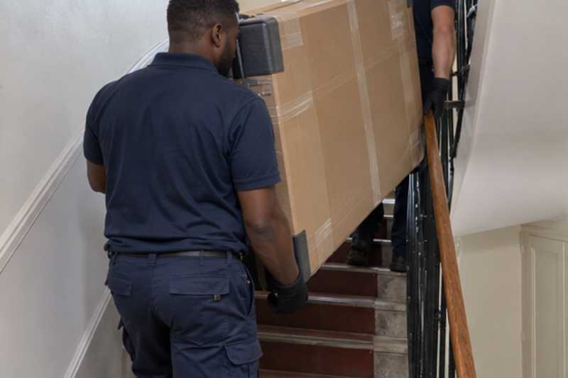 Deux déménageurs portent un grand téléviseur protégé par un carton épais, escalier étroit d'immeuble