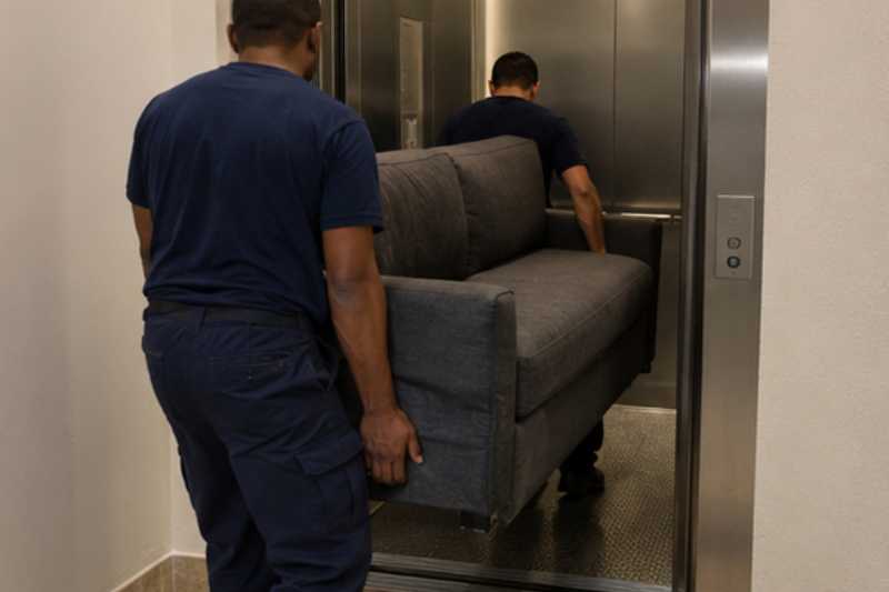 Deux déménageurs manœuvrent un canapé convertible lourd, palier devant un ascenseur étroit