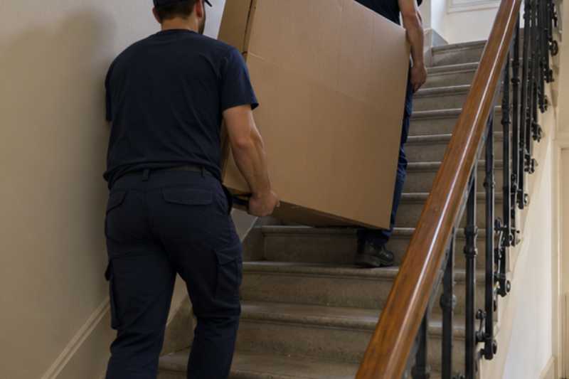 Deux déménageurs portent un grand téléviseur protégé par un carton épais, escalier d'immeuble sans ascenseur