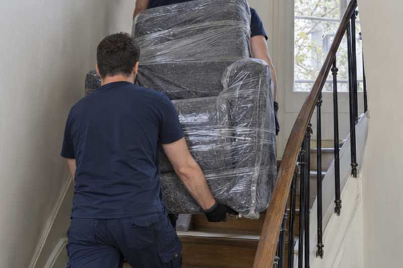 Deux déménageurs portent un fauteuil releveur emballé, escalier étroit d'immeuble