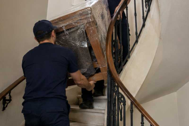 Déménageurs qui chargent une table en bois et six chaises emballées, escalier tournant avec un palier étroit