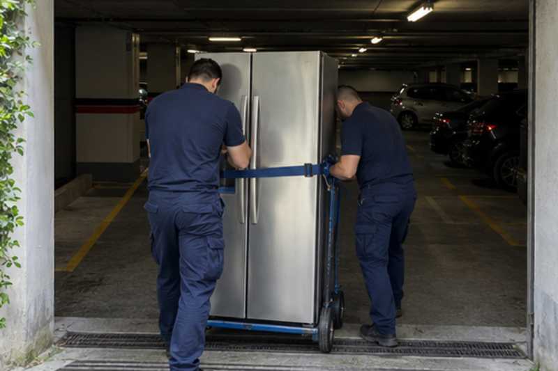 Deux déménageurs sanglent un réfrigérateur américain debout sur un diable, entrée de parking souterrain à hauteur limitée