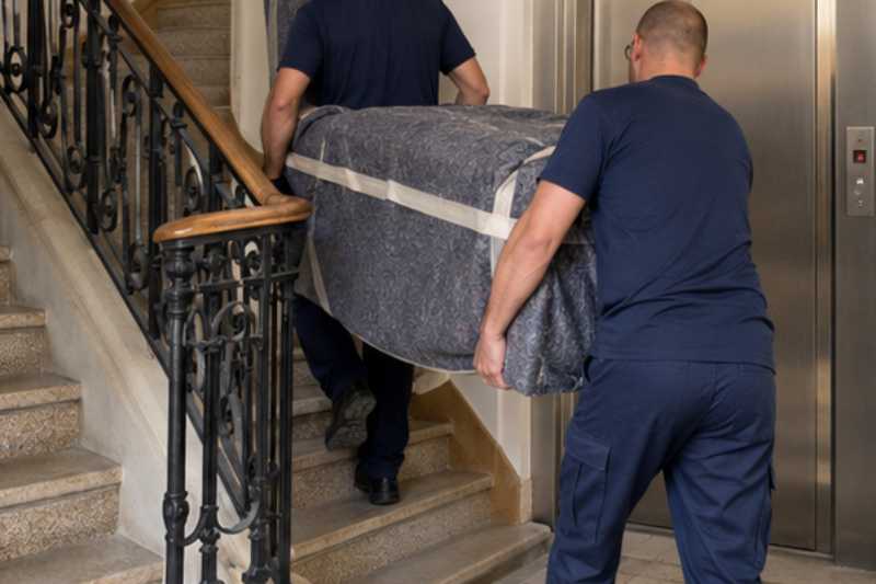Deux déménageurs portent des meubles emballés sous couvertures, cage d'escalier à côté d'un petit ascenseur