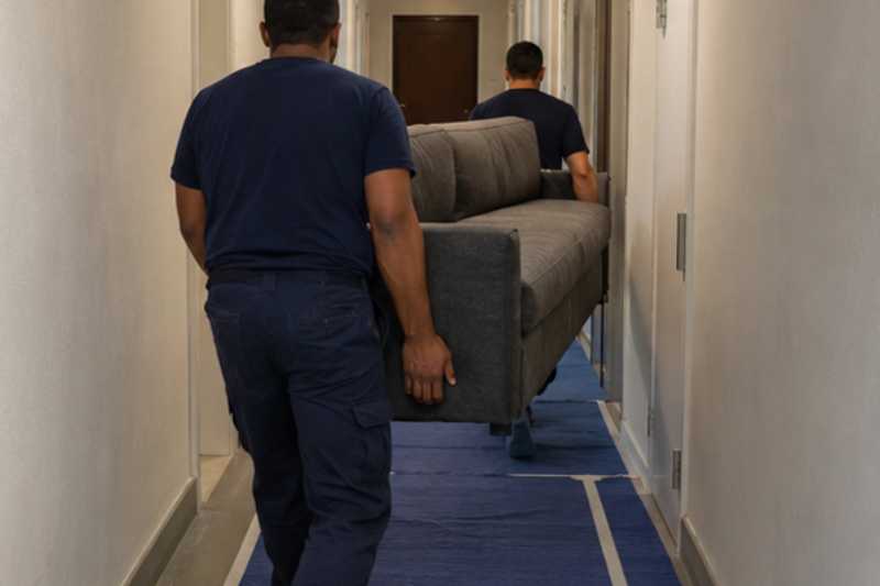 Deux déménageurs manœuvrent un canapé convertible lourd, long couloir d'immeuble protégé par des tapis