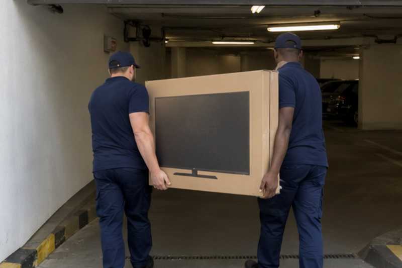 Deux déménageurs portent un grand téléviseur protégé par un carton épais, entrée de parking souterrain à hauteur limitée