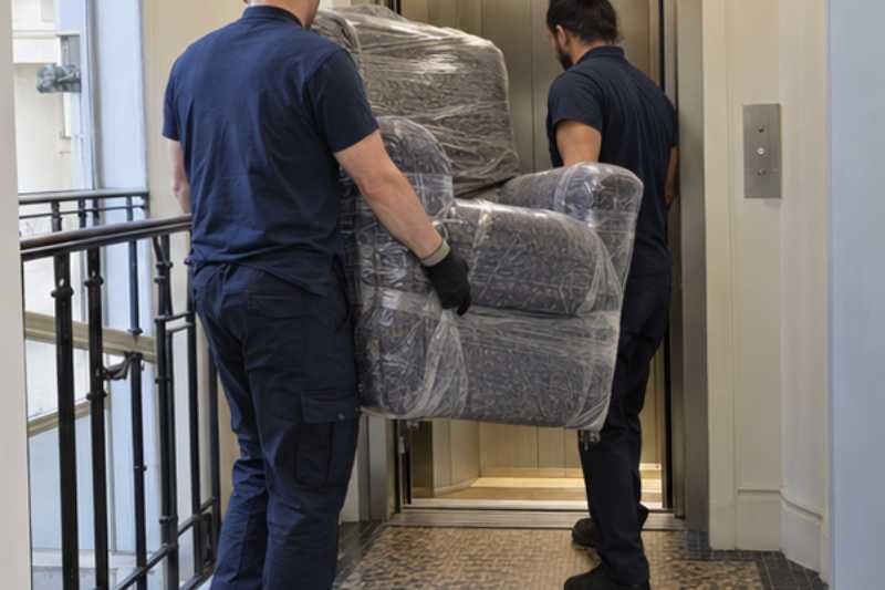 Deux déménageurs portent un fauteuil releveur emballé, palier devant un ascenseur étroit