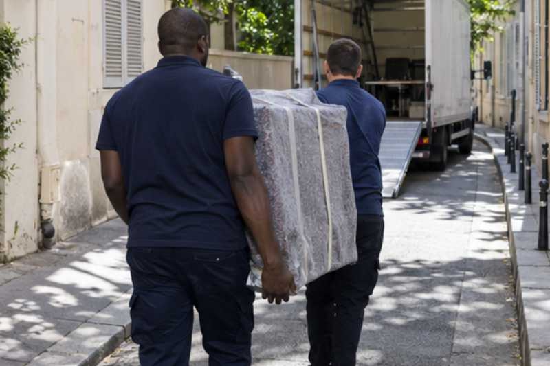 Deux déménageurs portent des meubles emballés sous couvertures, rue étroite, camion blanc garé plus loin
