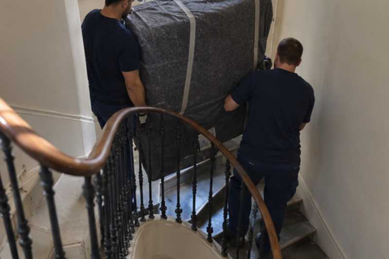 Deux déménageurs portent des meubles emballés sous couvertures, escalier tournant avec un palier étroit
