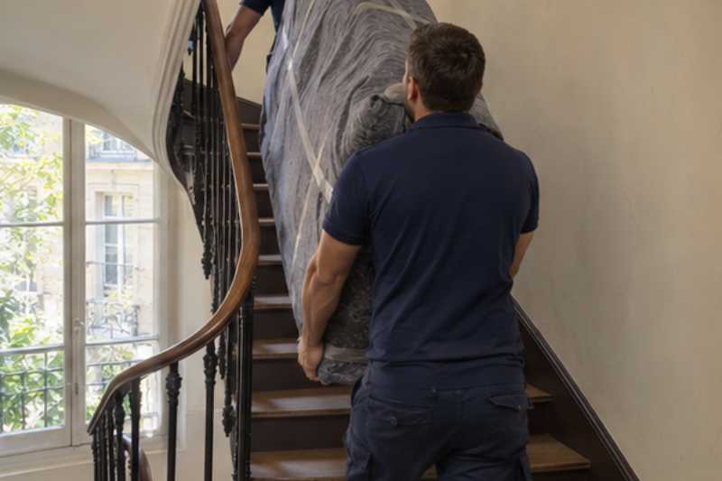 Deux déménageurs portent des meubles emballés sous couvertures, escalier d'immeuble sans ascenseur