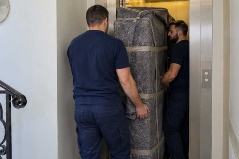Deux déménageurs portent des meubles emballés sous couvertures, palier devant un ascenseur étroit