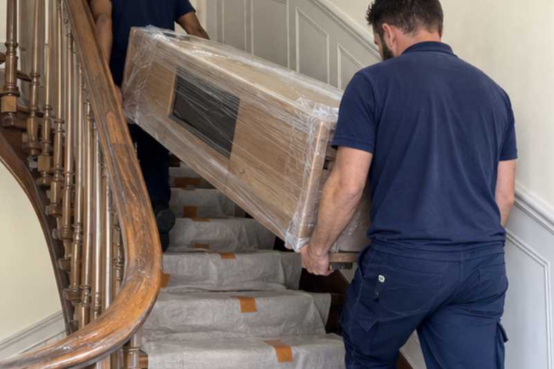 Déménageur qui porte un meuble TV bas entouré de film, escalier ancien en bois aux marches couvertes de protections